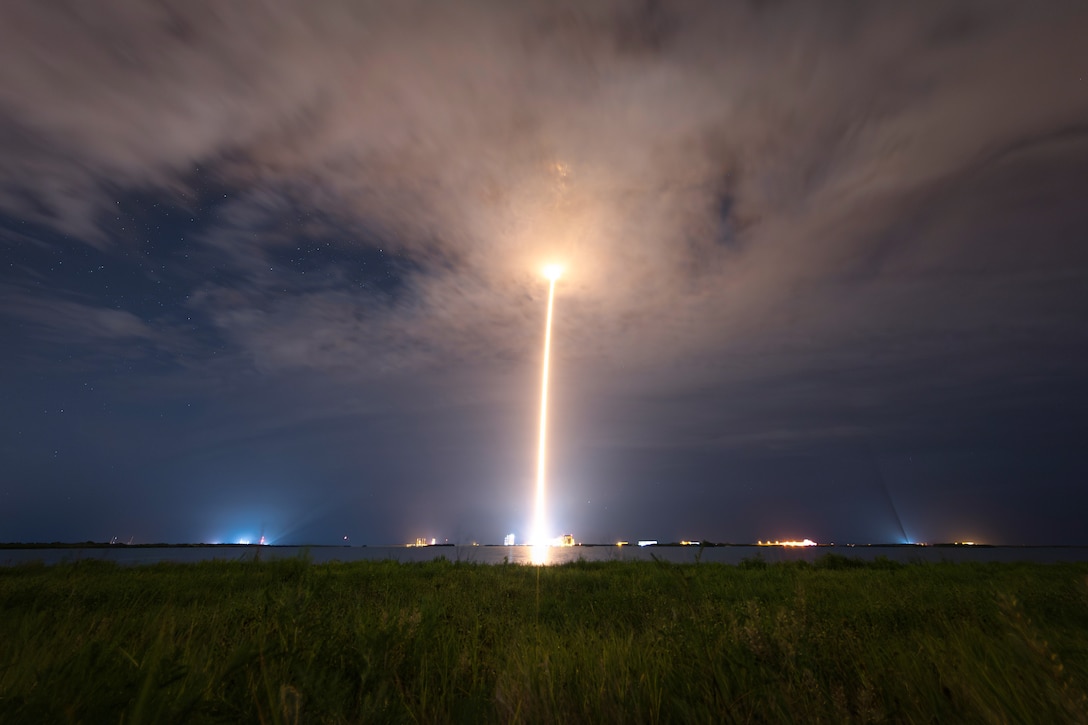 A rocket leaves a yellowish streak from a cloudy dark sky to field with lights in the background.