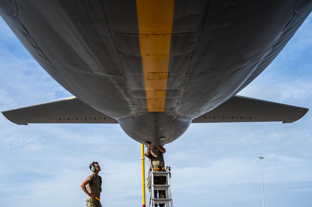An airman on a ladder works on the back upside of an aircraft as fellow airman watches from below against a blue sky.