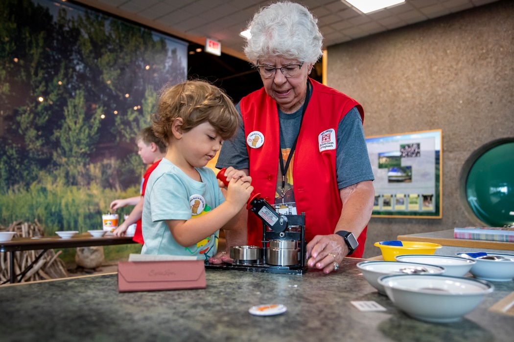 Linda Sunby, a volunteer at the U.S. Army Corps of Engineers’ Fort Peck Interpretive Center and Museum in Fort Peck, Montana, helps a young visitor create a commemorative button, June 21, 2025, during the center’s 20th anniversary celebration.
