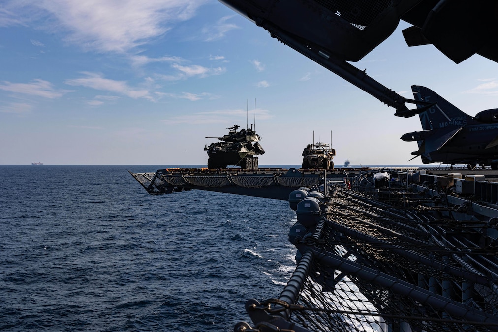 Two armored vehicles ride on the side of a ship near nets as another ship sails in the distance.