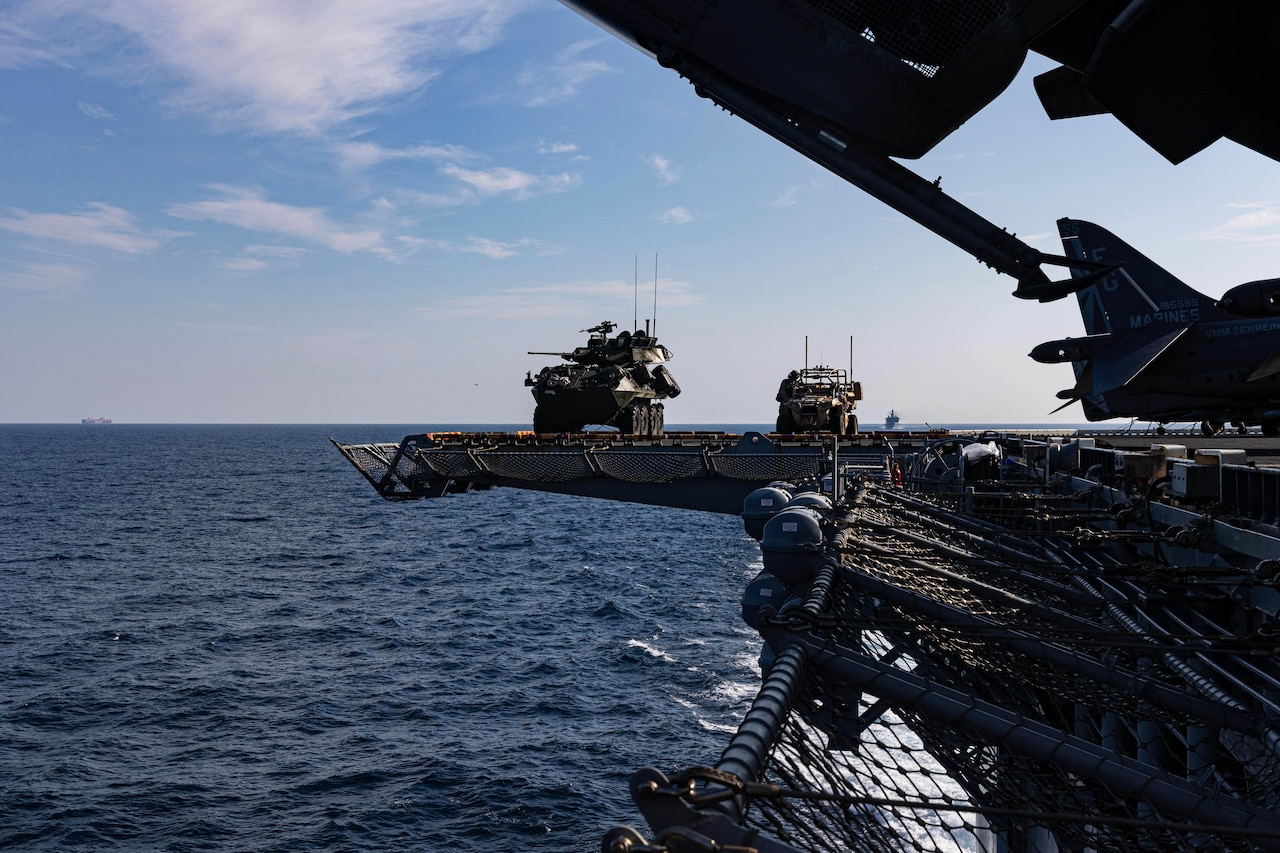 Two armored vehicles ride on the side of a ship near nets as another ship sails in the distance.