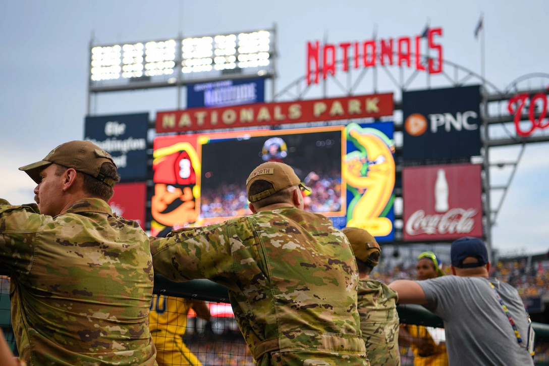 Service members watch a baseball game from the dugout.