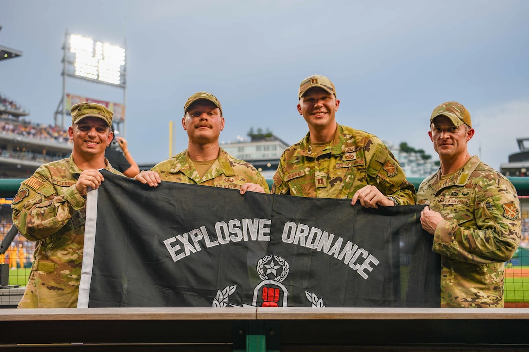 Service members hold up a flag in the dugout of a baseball stadium.