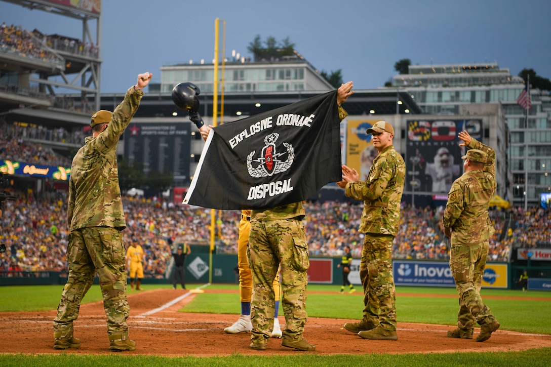 Service members wave a flag on a baseball field.