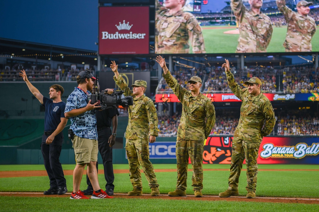 Service members wave on a baseball field.