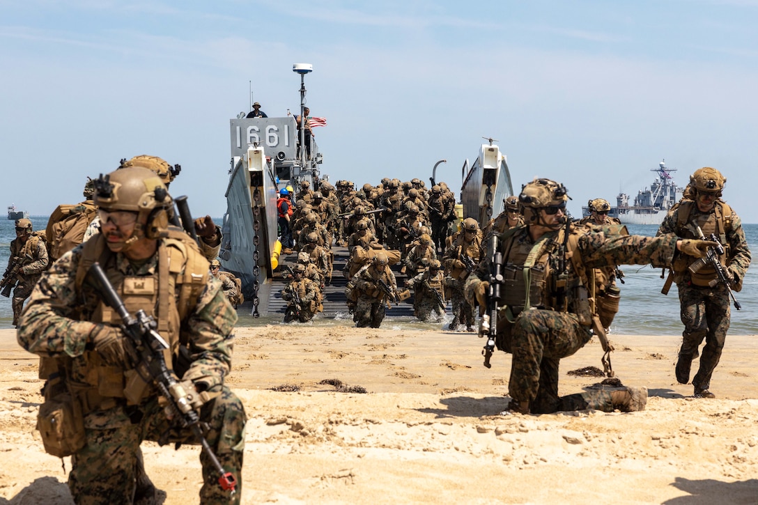 Marines kneel in tactical gear while holding weapons on a beach as fellow Marines unload from a ship in the background.