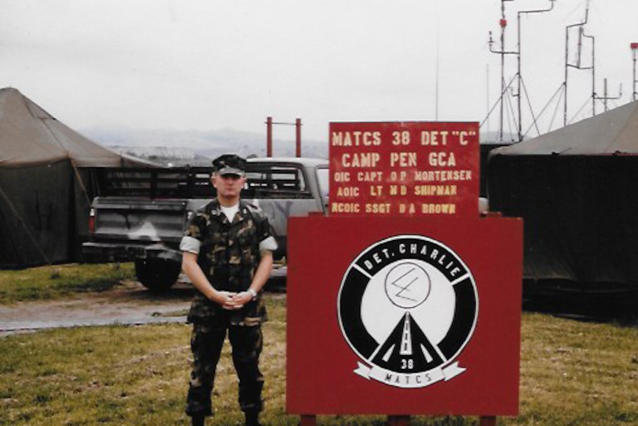 A man dressed in a camouflage uniform stands beside a sign with tents in the background.