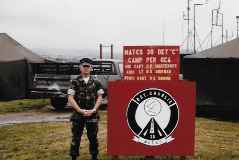 A man dressed in a camouflage uniform stands beside a sign with tents in the background.