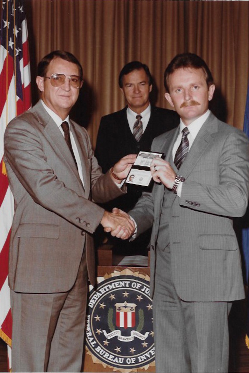 Two men in business attire shake hands as another man smiles behind them. The man on the left is handing the man on the right an FBI badge.