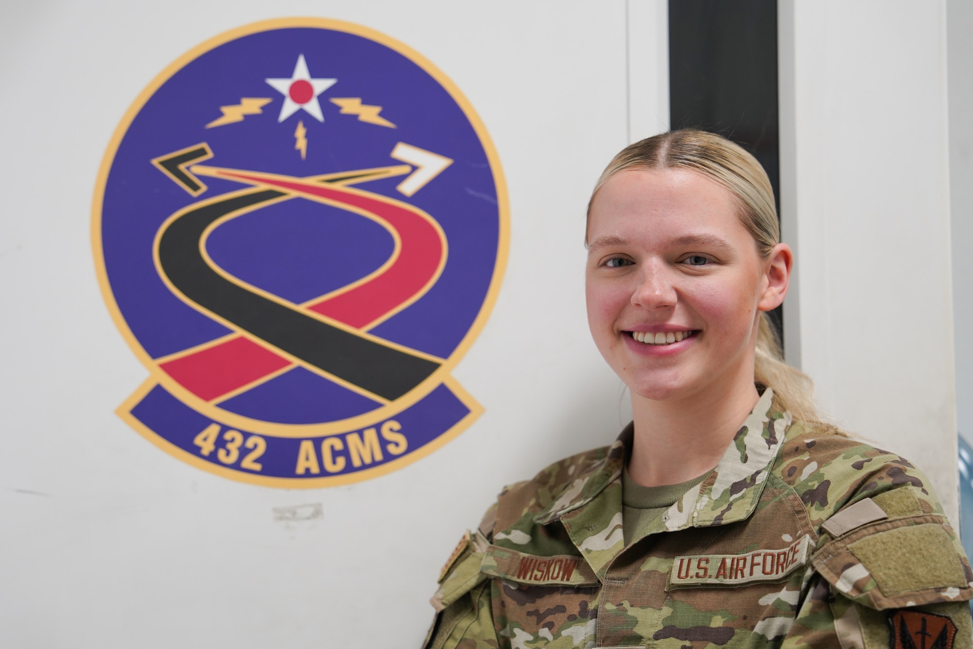 A woman in a military uniform poses in front of a patch for the 432 ACMS.
