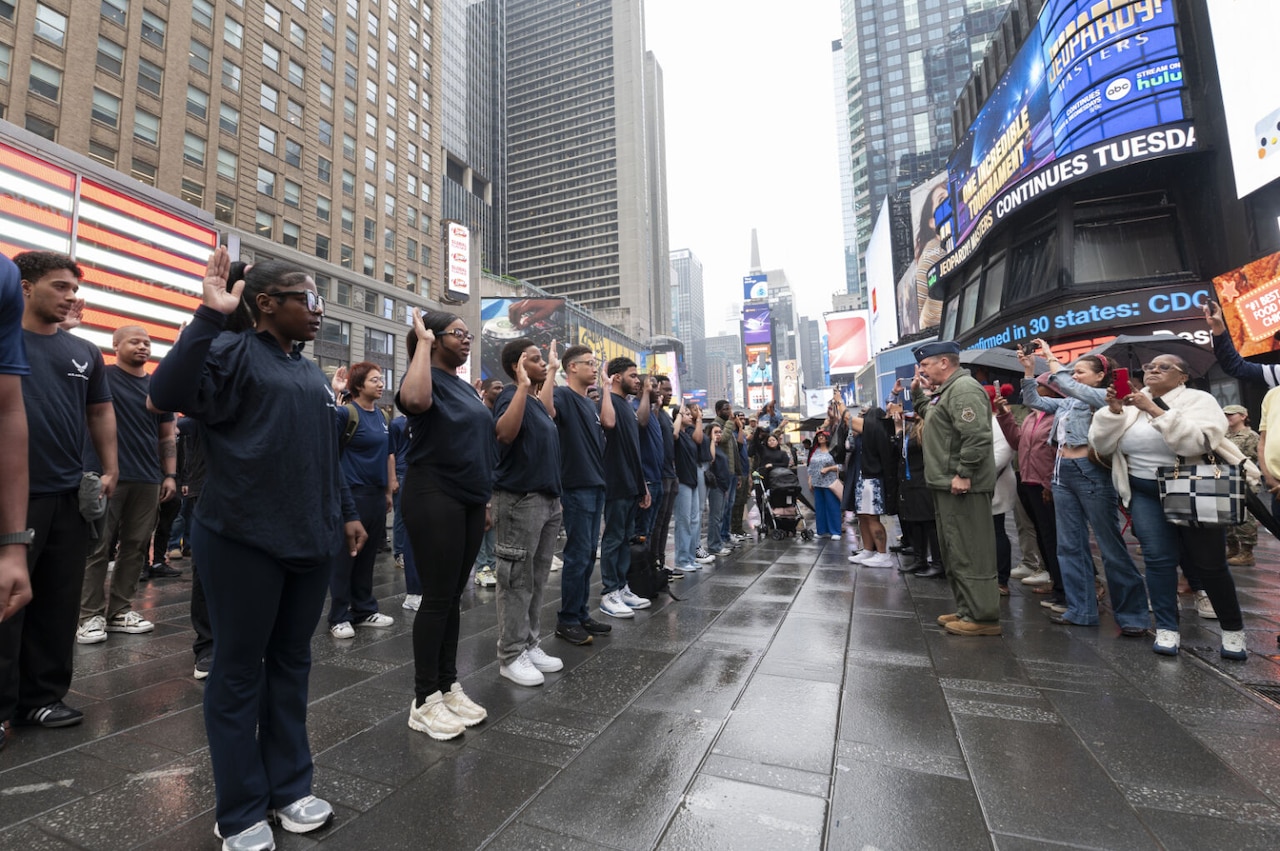 Amidst tall buildings, some with electronic signs, more than a dozen young people dressed in blue shirts stand in a formation and raise their right hands. In front of them, a man dressed in a military uniform faces them while raising his right hand. Onlookers take photos.