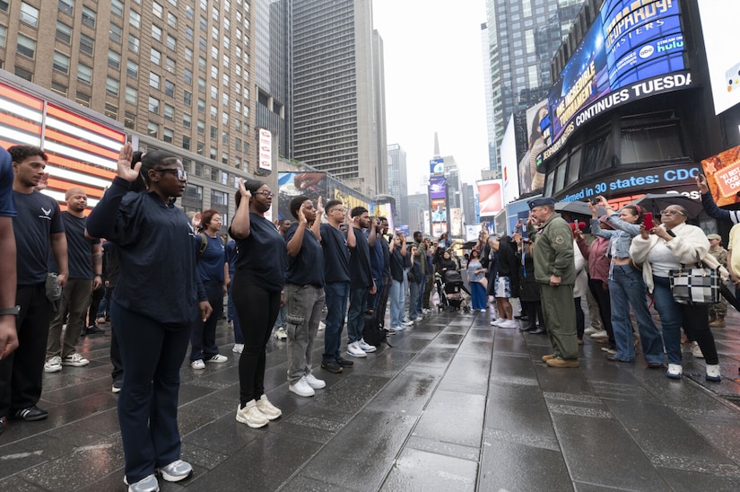 Amidst tall buildings, some with electronic signs, more than a dozen young people dressed in blue shirts stand in a formation and raise their right hands. In front of them, a man dressed in a military uniform faces them while raising his right hand. Onlookers take photos.