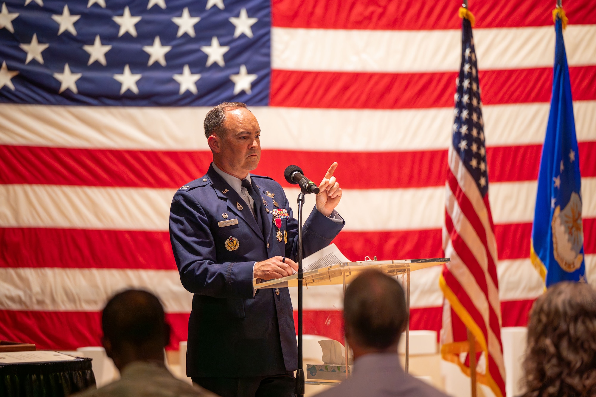 Shoemaker speaks at a podium with a flag in the background.
