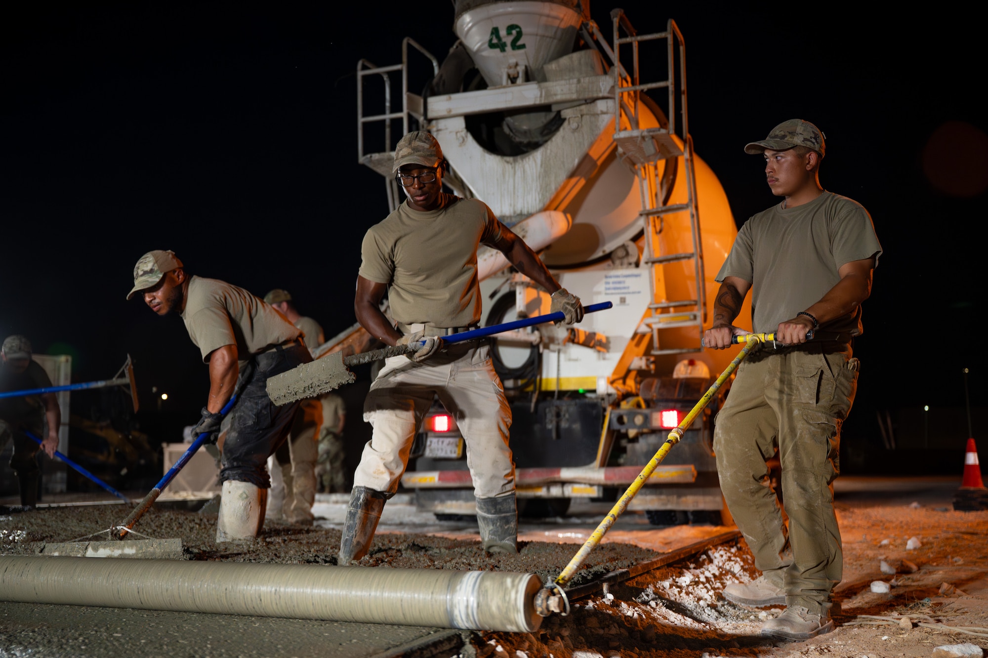 Three U.S. Air Force Airmen level a wet concrete pad at night.