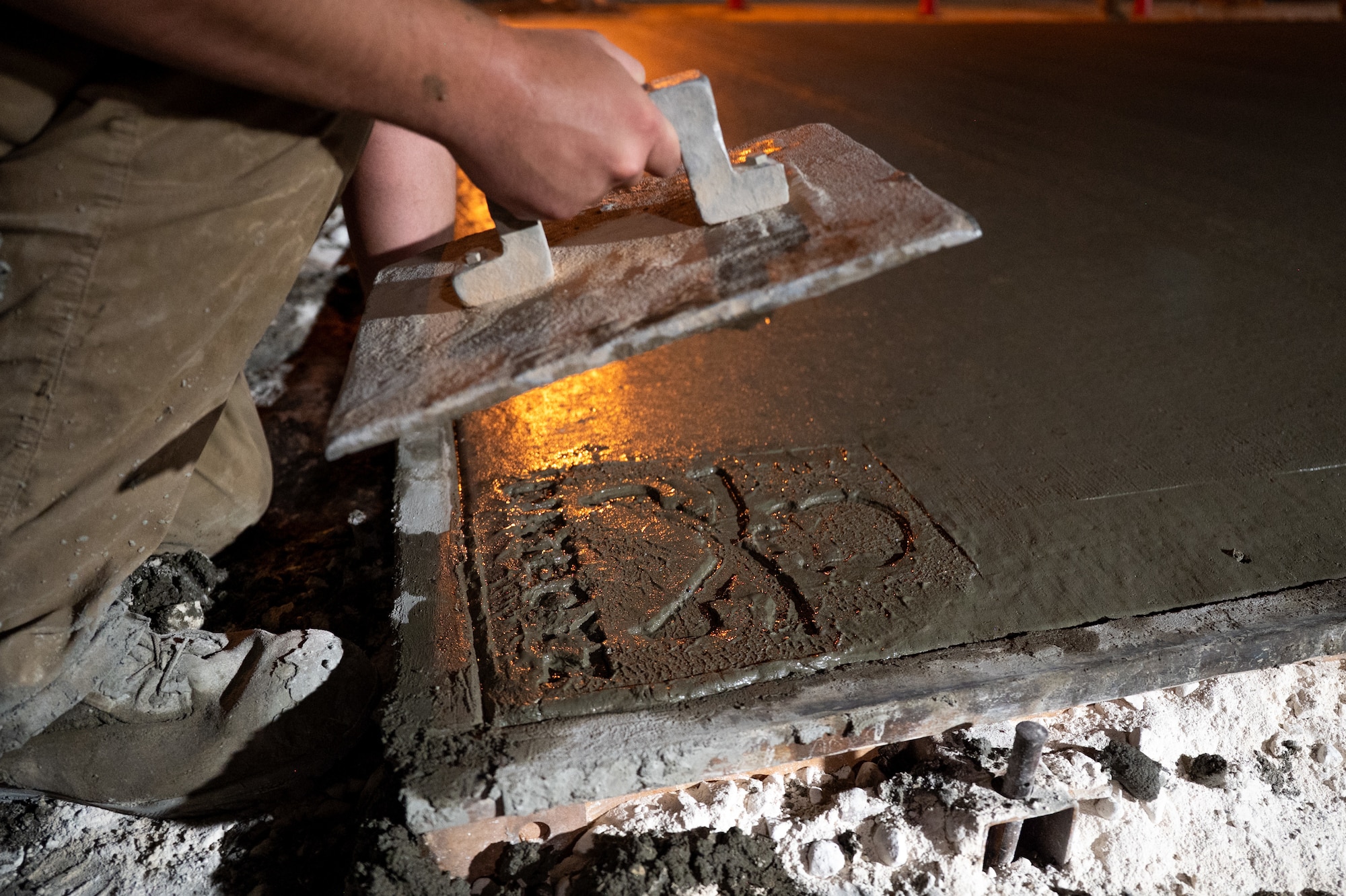 U.S. Air Force Airman pulls a concrete stamp with "DIRTBOYZ" from a wet concrete pad at night