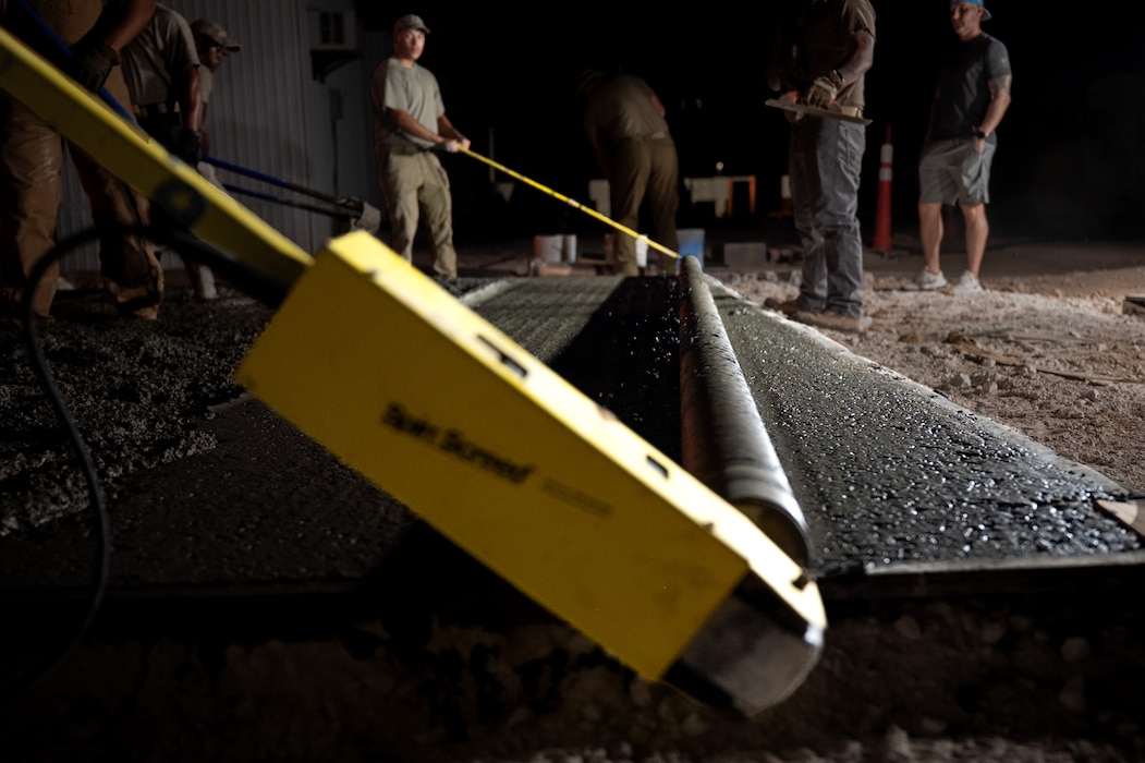 U.S. Air Force Airmen maneuver a roller screed along a wet concrete pad at night.