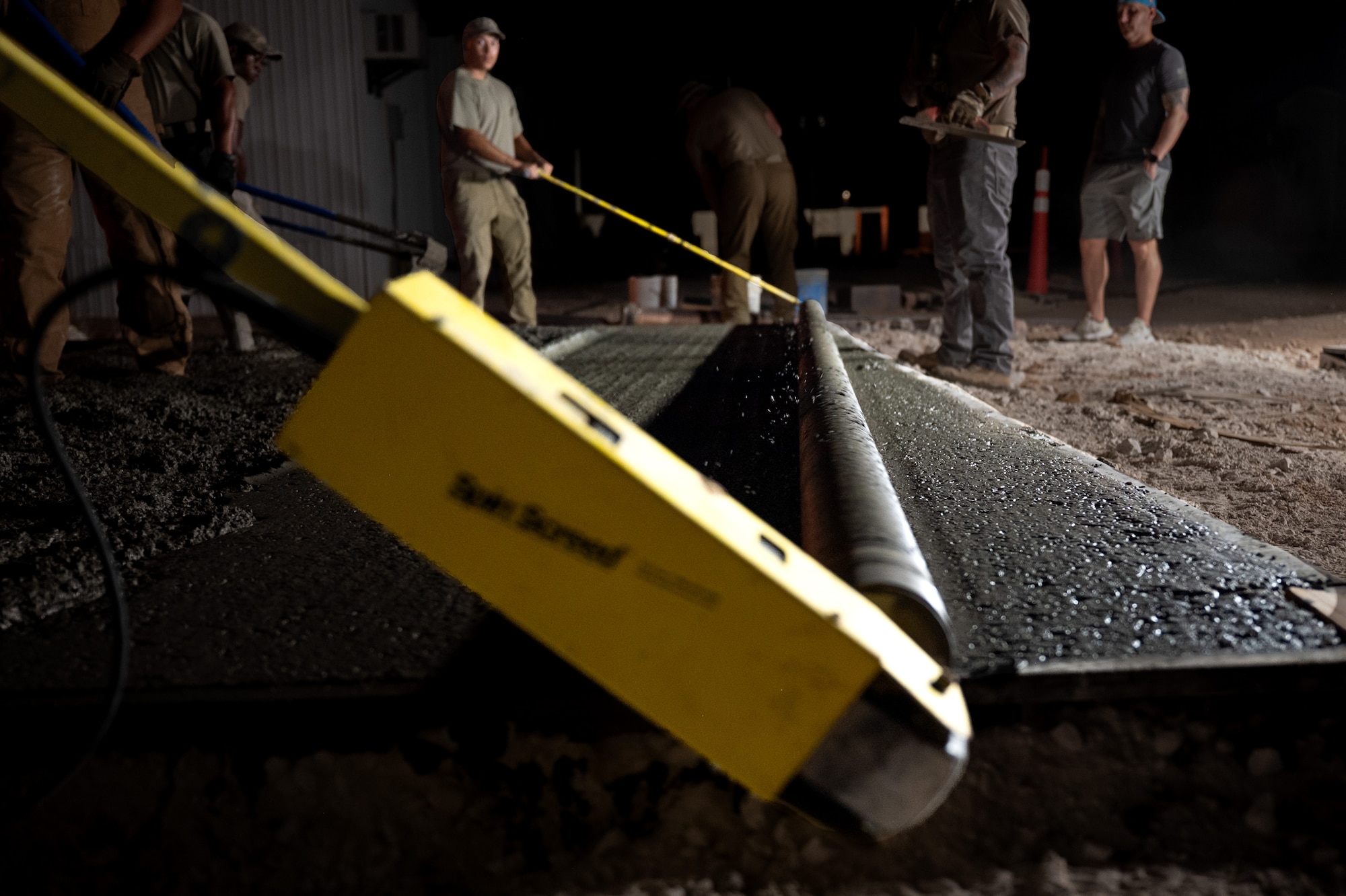 U.S. Air Force Airmen maneuver a roller screed along a wet concrete pad at night.