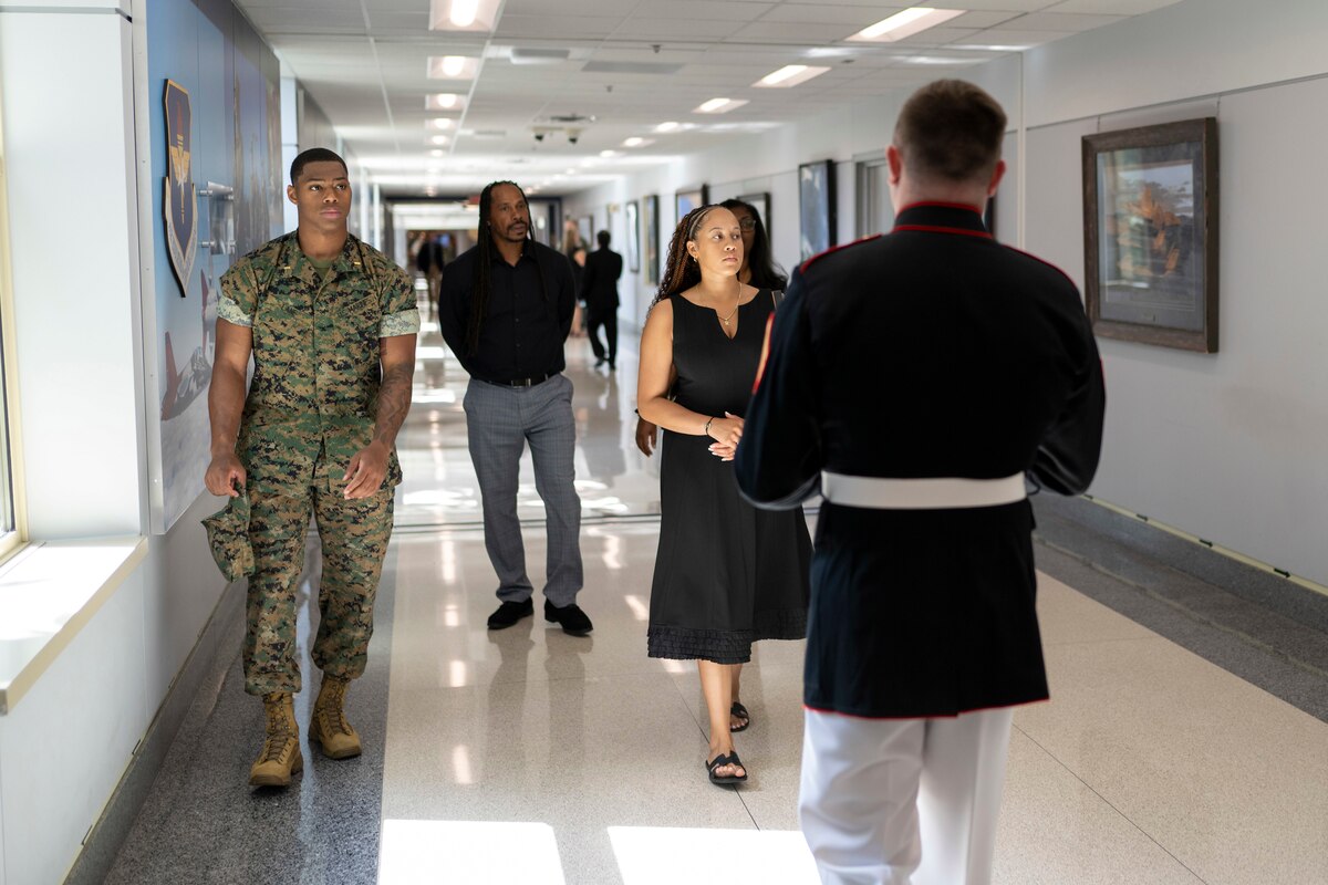A Marine and civilians walk down a hallway toward a service member facing them and walking backwards.