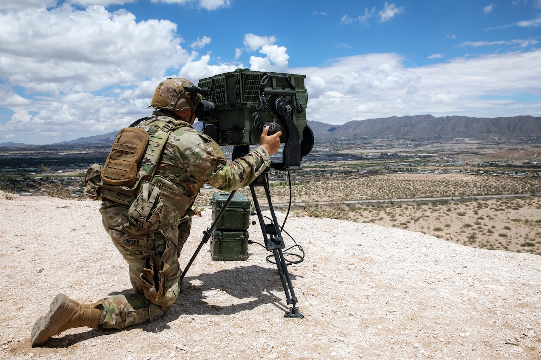 A soldier kneels on one knee to look through a surveillance device toward a sparsely vegetated area with mountains in the distance.