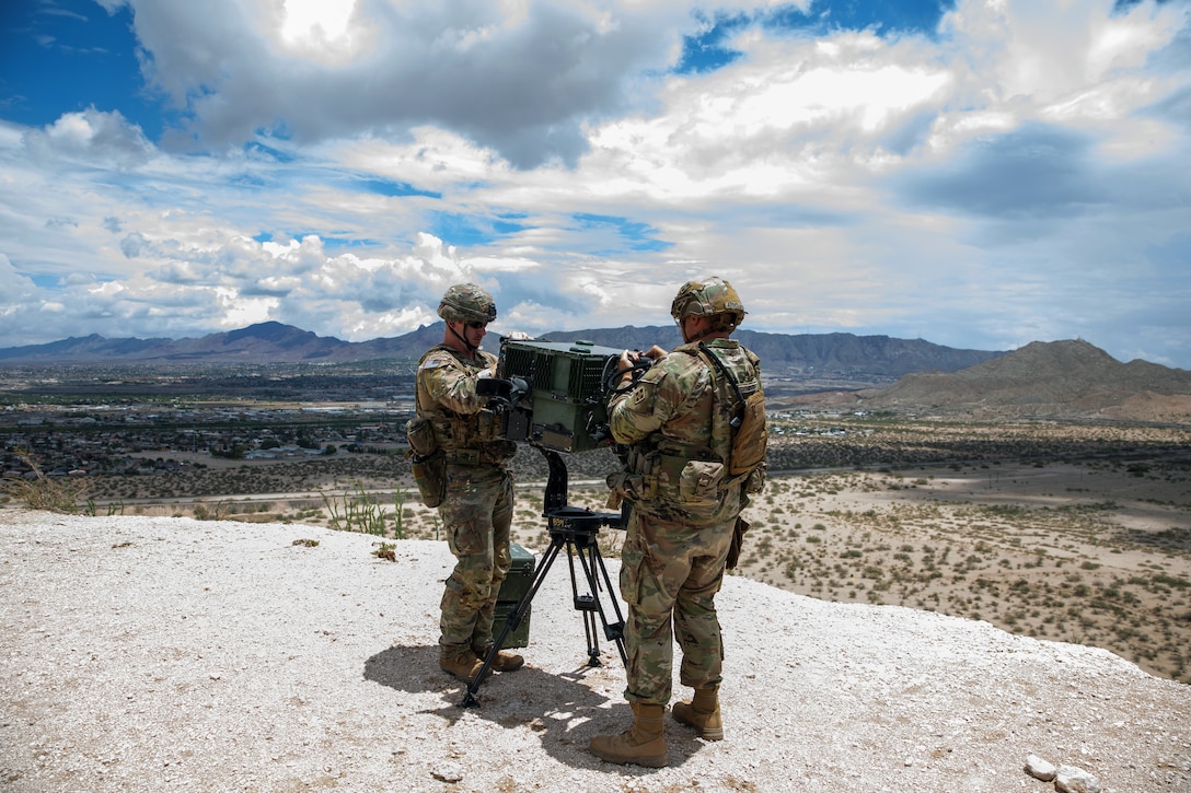Two soldiers work on a surveillance device from a gravelly, elevated area overlooking sparsely vegetated land with mountains in the distance.