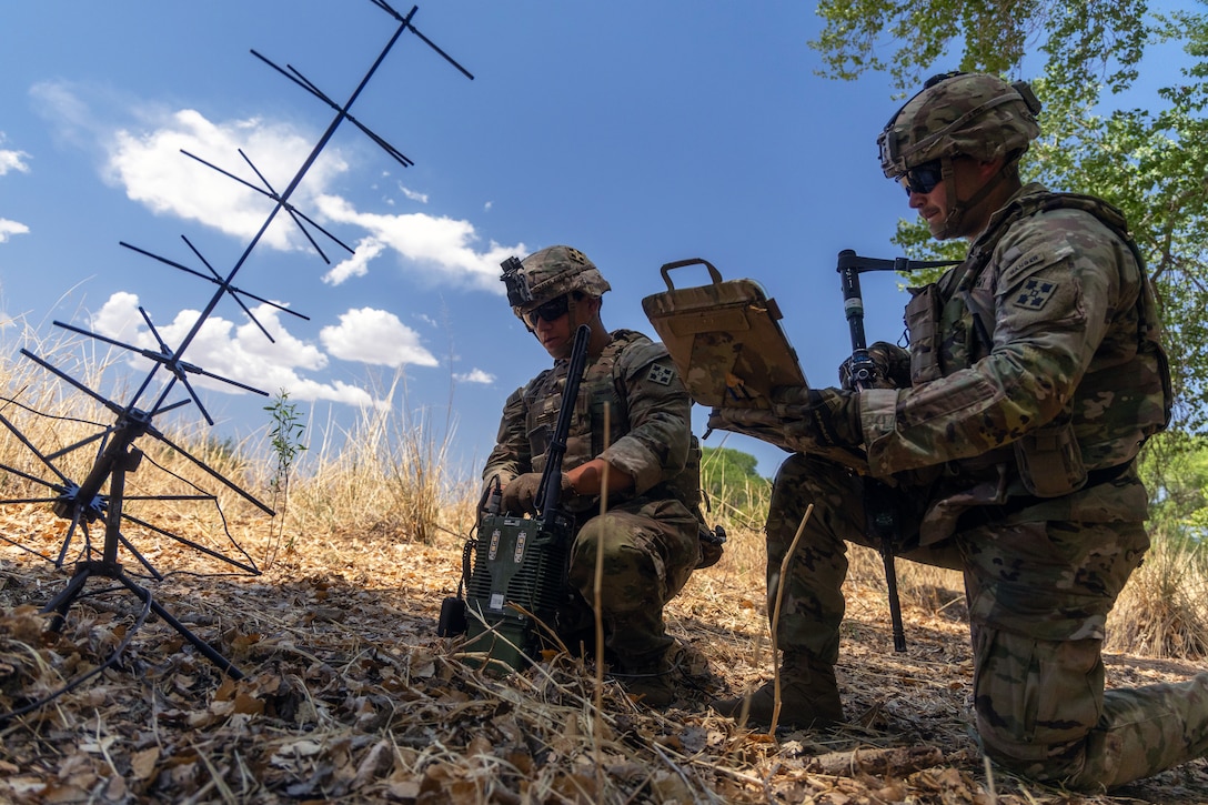 Two kneeling soldiers check communications devices near an antenna placed on the ground on a sunny day.