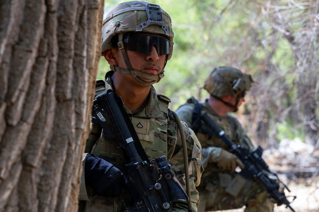 Two soldiers in combat gear, wearing sunglasses and holding rifles kneel and check their surroundings in a wooded area.