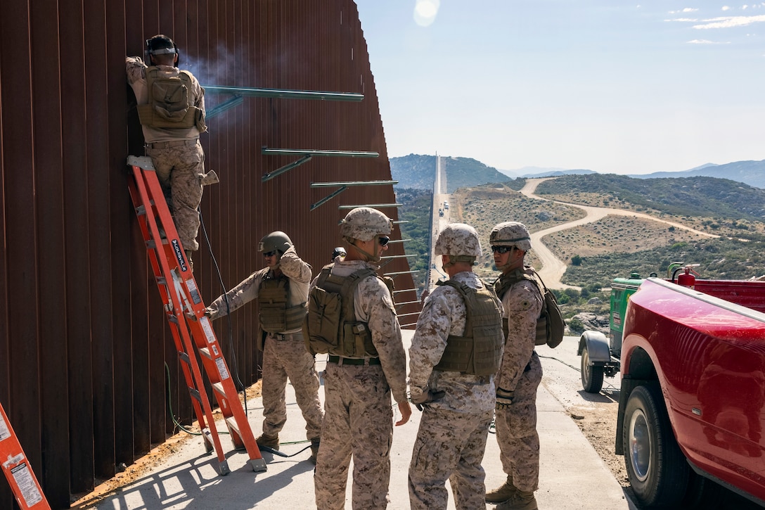 Marines, one standing on a ladder, install additional security obstacles to a tall wall along a road on a sunny day.