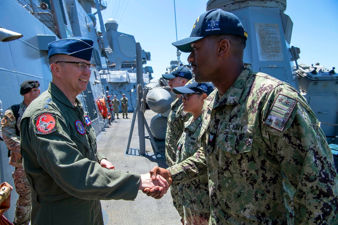 An airman shakes hands with a sailor standing next to two other sailors on a ship's deck on a sunny day; others stand in the background.