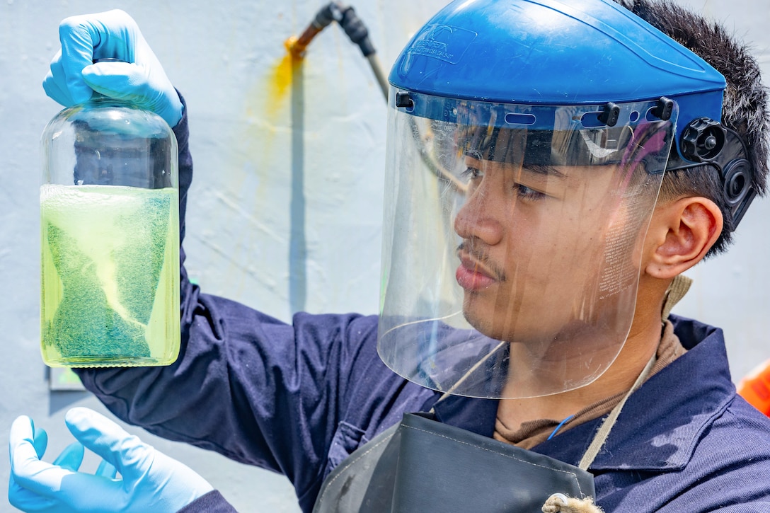 A close-up of a sailor wearing face shield and gloves holding a fuel sample.