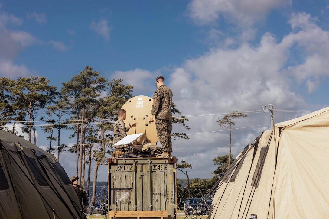 Two Marines work on a satellite while standing on a wood box in between two tents under a blue sky.