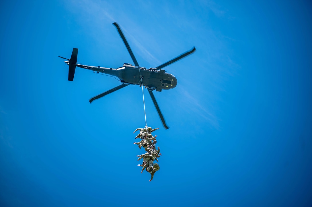 A dozen soldiers hang from a rope connected to a hovering helicopter against a blue sky as seen from below.