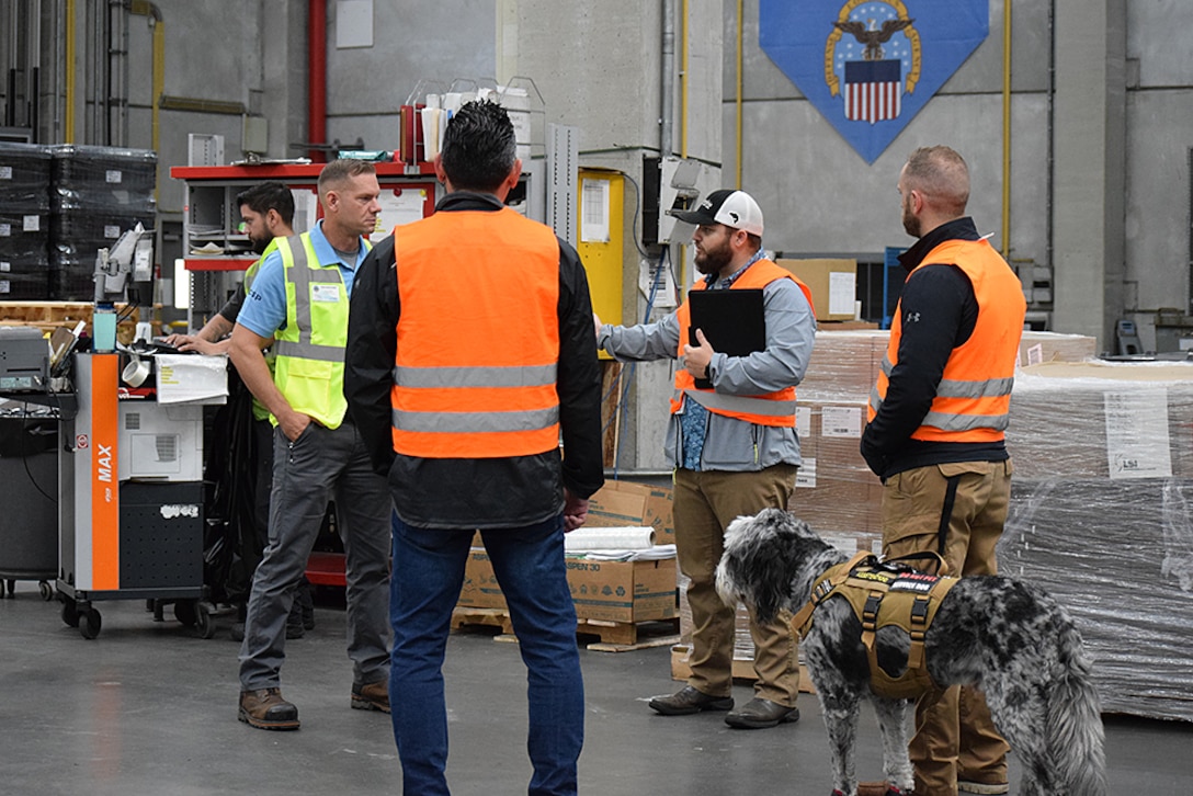 Men and a dog talk in a warehouse.