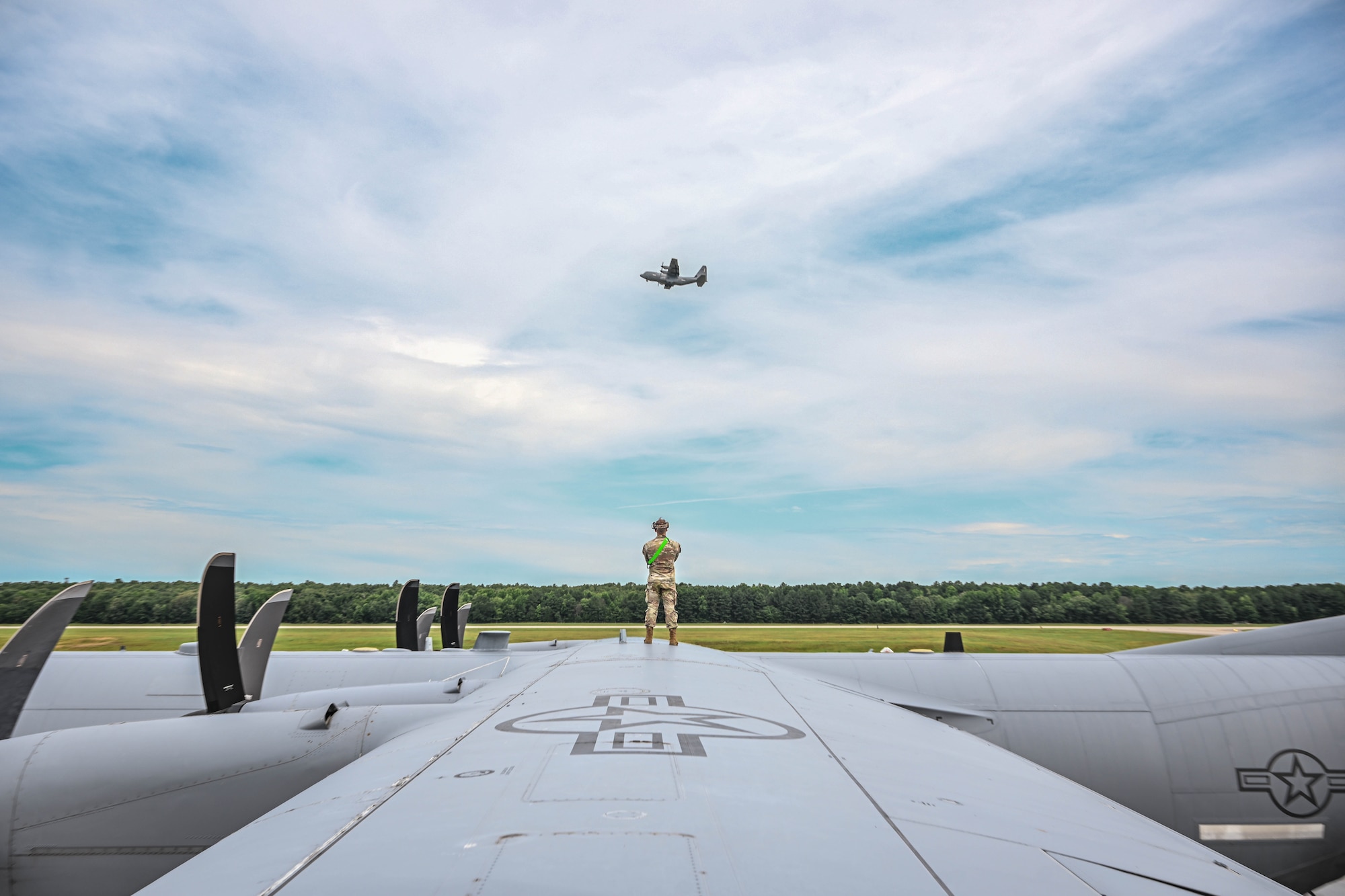 A man in uniform stands on top of an aircraft as he watches another aircraft takeoff.