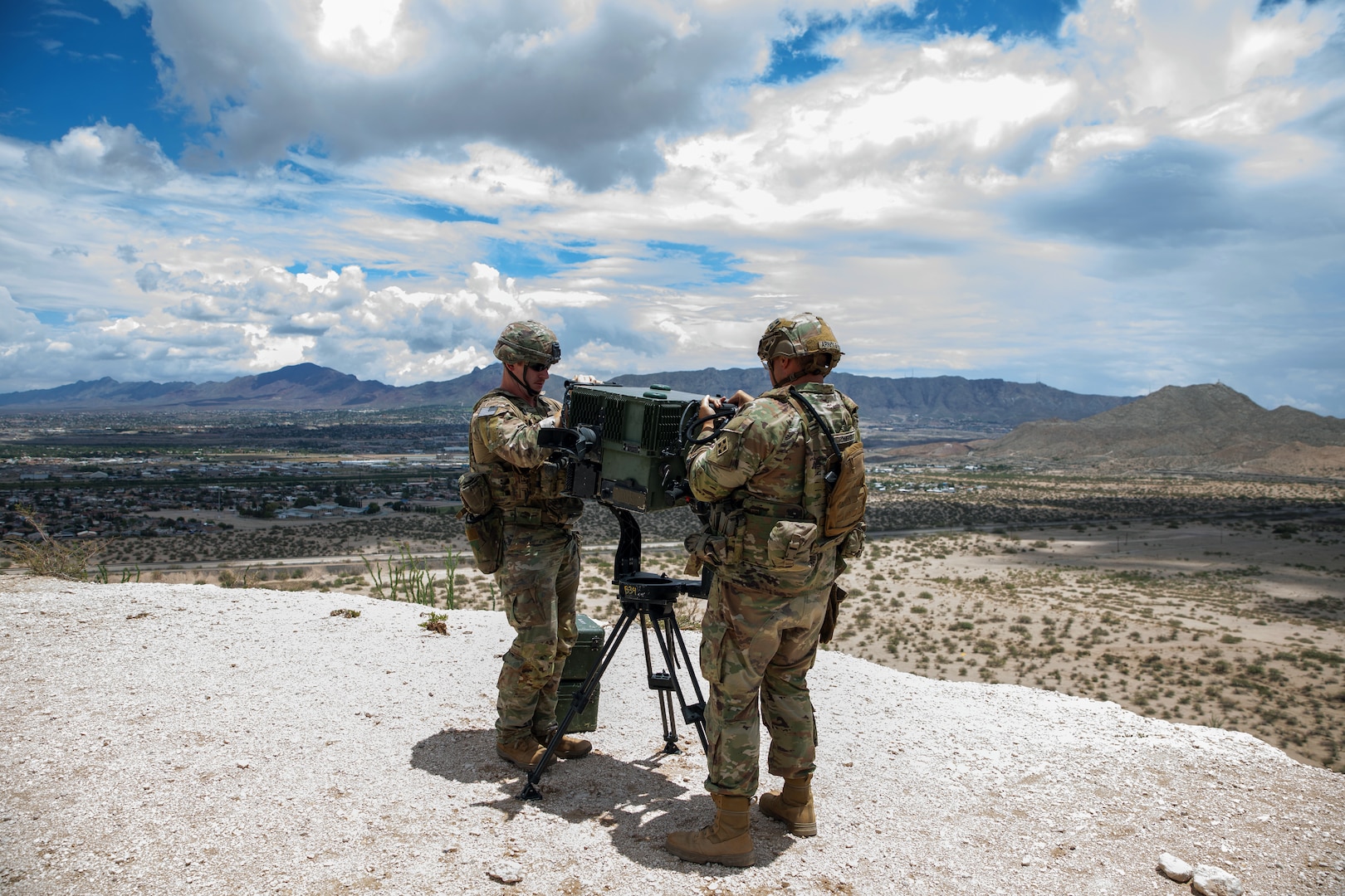 Two soldiers work on a surveillance device from a gravelly, elevated area overlooking sparsely vegetated land with mountains in the distance.