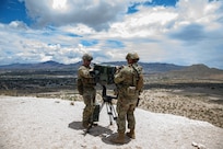 Two soldiers work on a surveillance device from a gravelly, elevated area overlooking sparsely vegetated land with mountains in the distance.