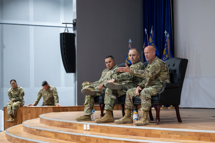 A group of three senior leaders seated at the center edge of a stage speaks to a crowd out of frame