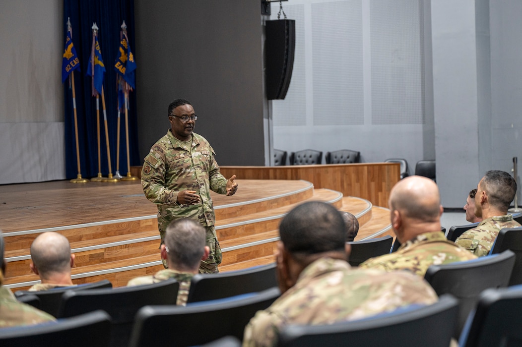 Master Sgt. Corker speaks from the left of frame to a crowd along the entire foreground with a stage behind him