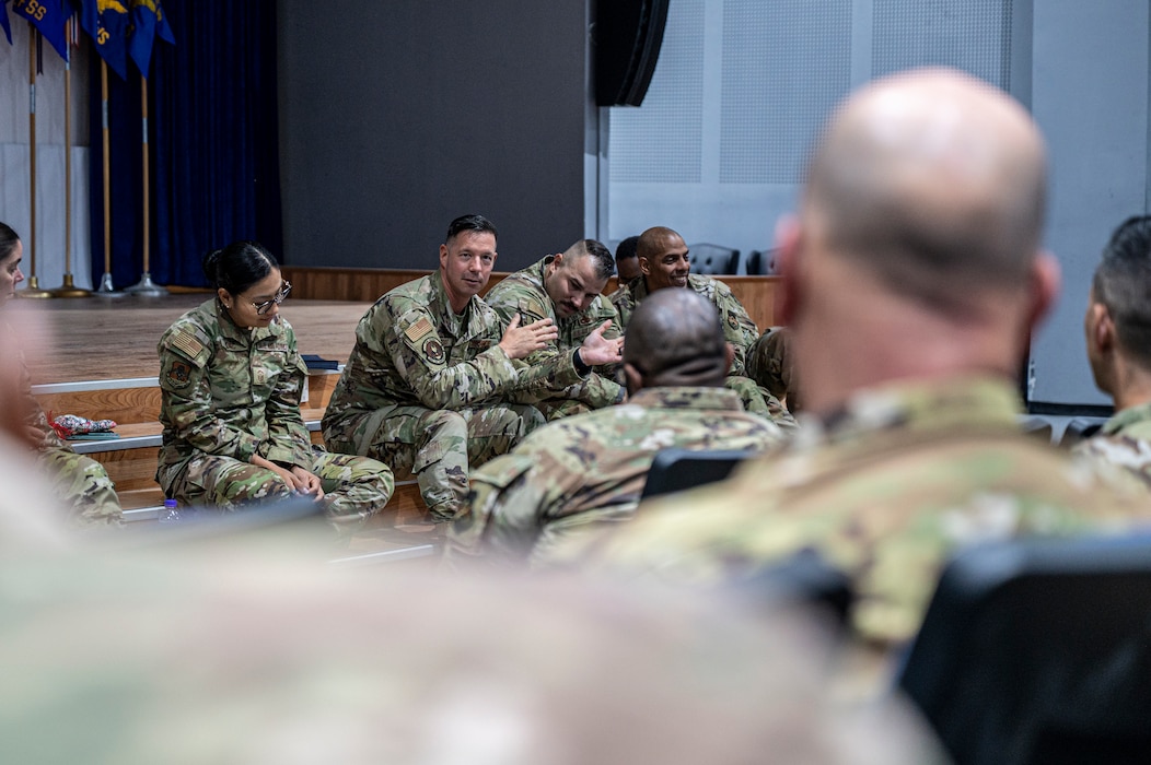 A lineup of first sergeants sits at the stairs of a stage in the background, with Master Sgt. Brooks in the middle speaking to the crowd in the foreground