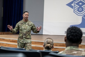 Master Sgt. Brooks speaks with his hands out at the left of frame to a crowd in the foreground with a stage behind him