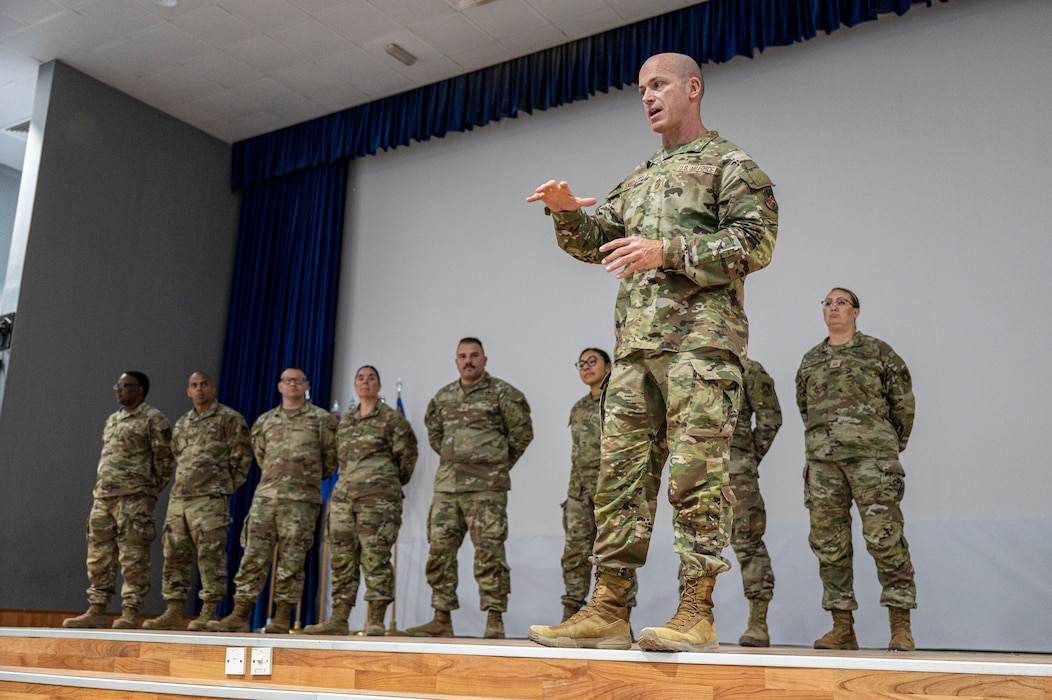 Chief Ski speaks in the foreground of a stage to a crowd out of frame with a lineup of first sergeants behind him at parade rest