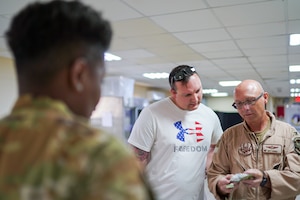 Col. Grimes at the right of frame shows the contents of a JFAK to Tech. Sgt. Campanella, who is out of uniform on his off-hours, as he looks over the shoulder to the showcase, with another Airman in the foreground out of focus