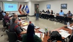 A man stand near the center of a class room surrounded by tables with students seated at them looking towards the man standing. it appears that the man is teaching the students.