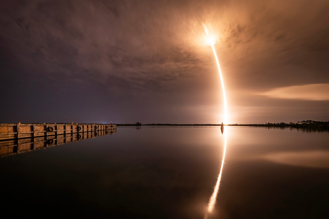 A rocket launches leaving a yellowish streak from a cloudy dark sky to a body of water as its reflection is seen in the foreground.
