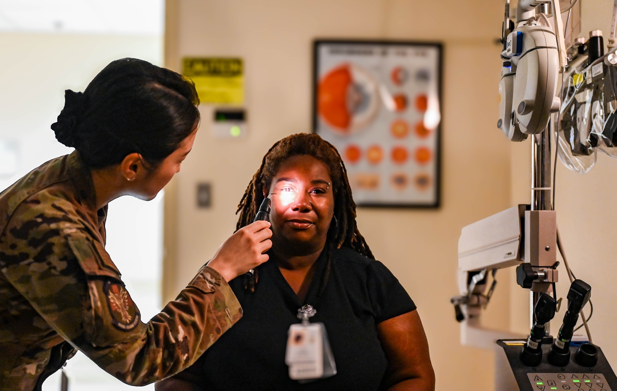 A military optometrist conducts a pupil exam on a woman in civilian clothing by shining a light into her eye.