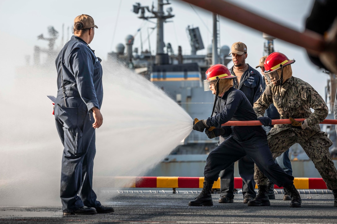 Sailors wearing firefighting helmets spray a water hose aboard a ship as fellow sailors watch.