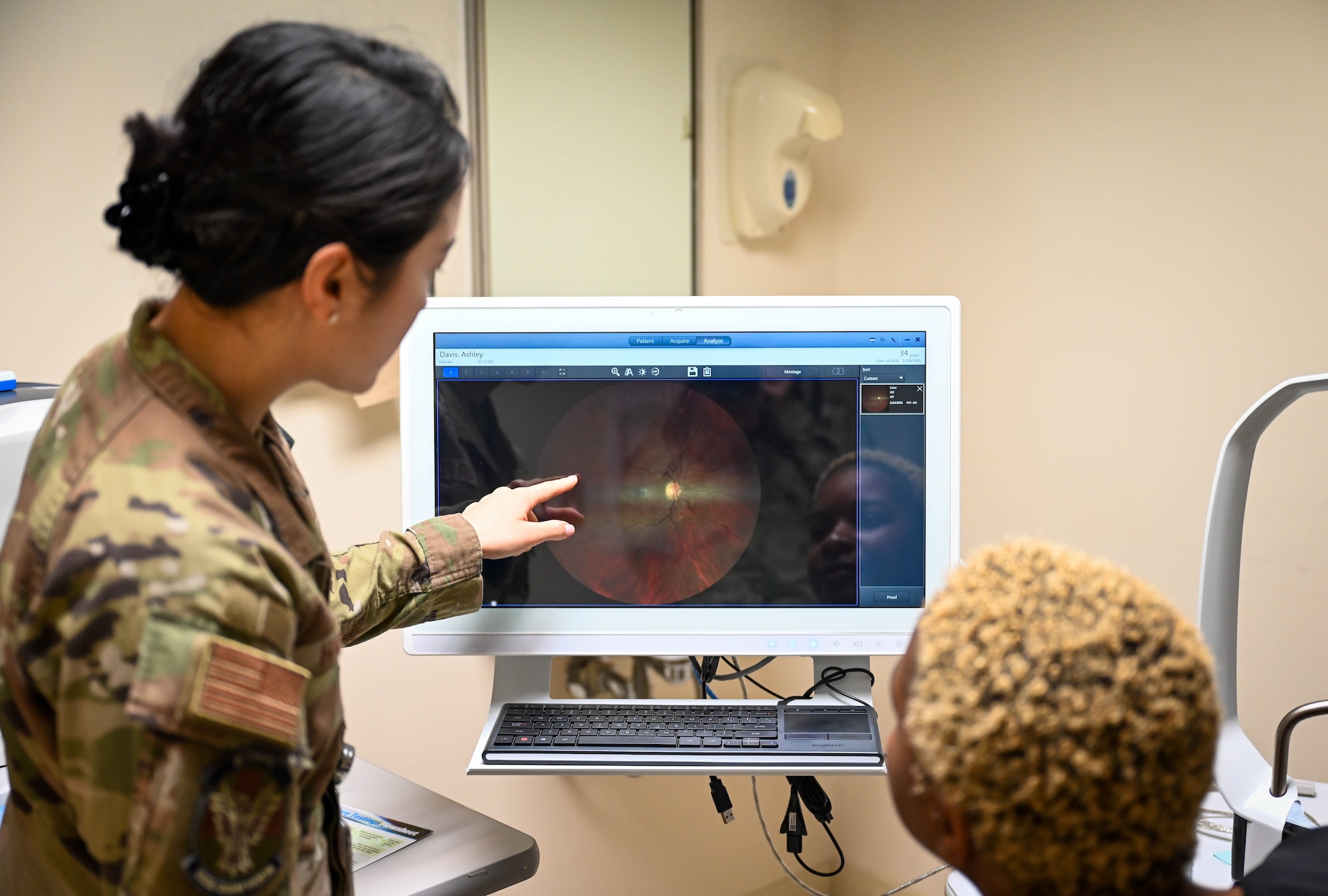 A woman in military uniform points to a screen showing eye scan results to a patient.