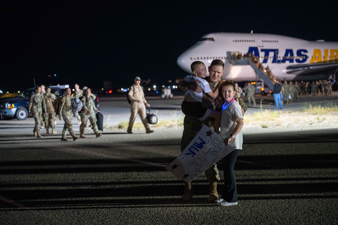 Two children, one holding a sign, hug an airman as fellow airmen carrying luggage exit a plane in the dark in the background.