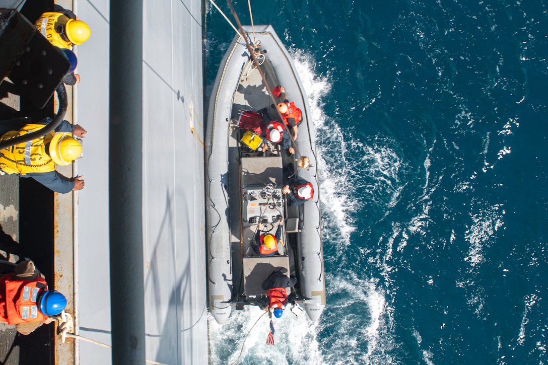 Sailors in a small boat are lowered into a body of water as fellow sailors watch from the side of a ship as seen from above.
