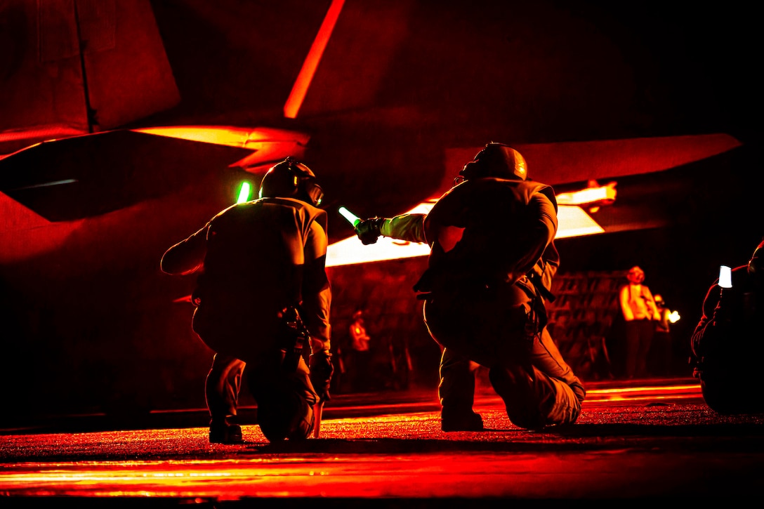 Two sailors holding neon batons kneel next to an aircraft aboard a ship in the dark illuminated by red light.
