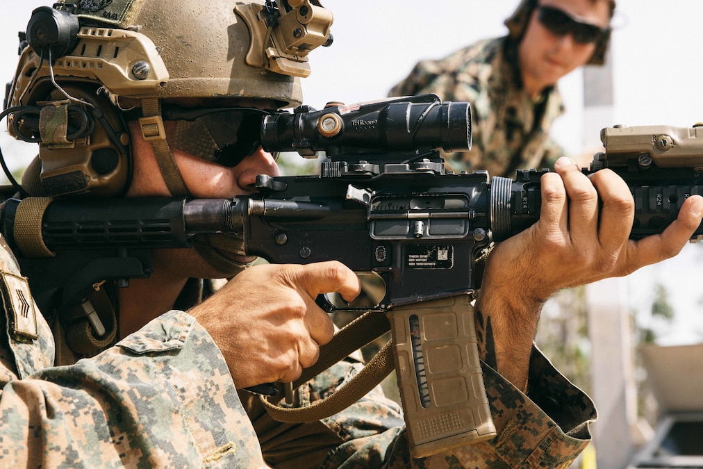 A close-up of a Marine in tactical gear firing a weapon as fellow service member watches in the blurred background.